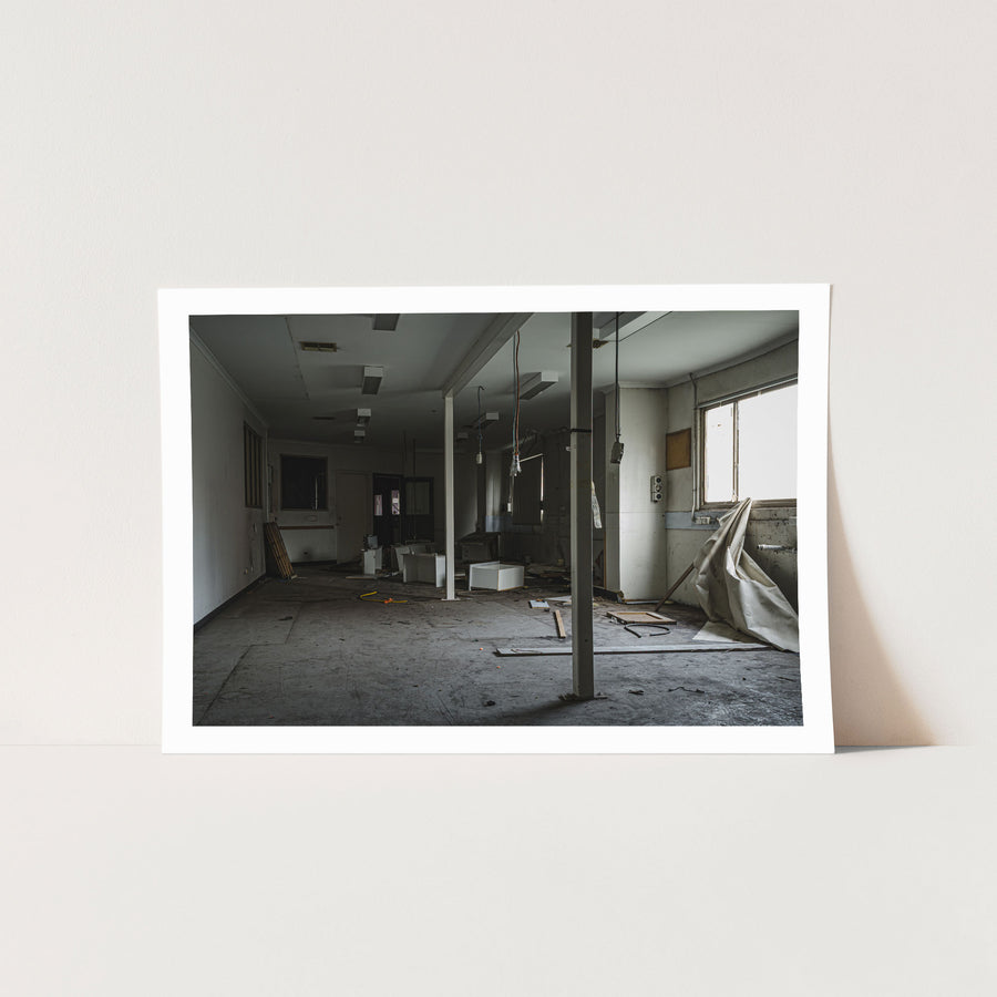 Stripped interior room at the former Bradmill factory in Yarraville, with a white support column, scattered cabinetry panels and cables on a bare concrete floor, and daylight entering from a window on the right.