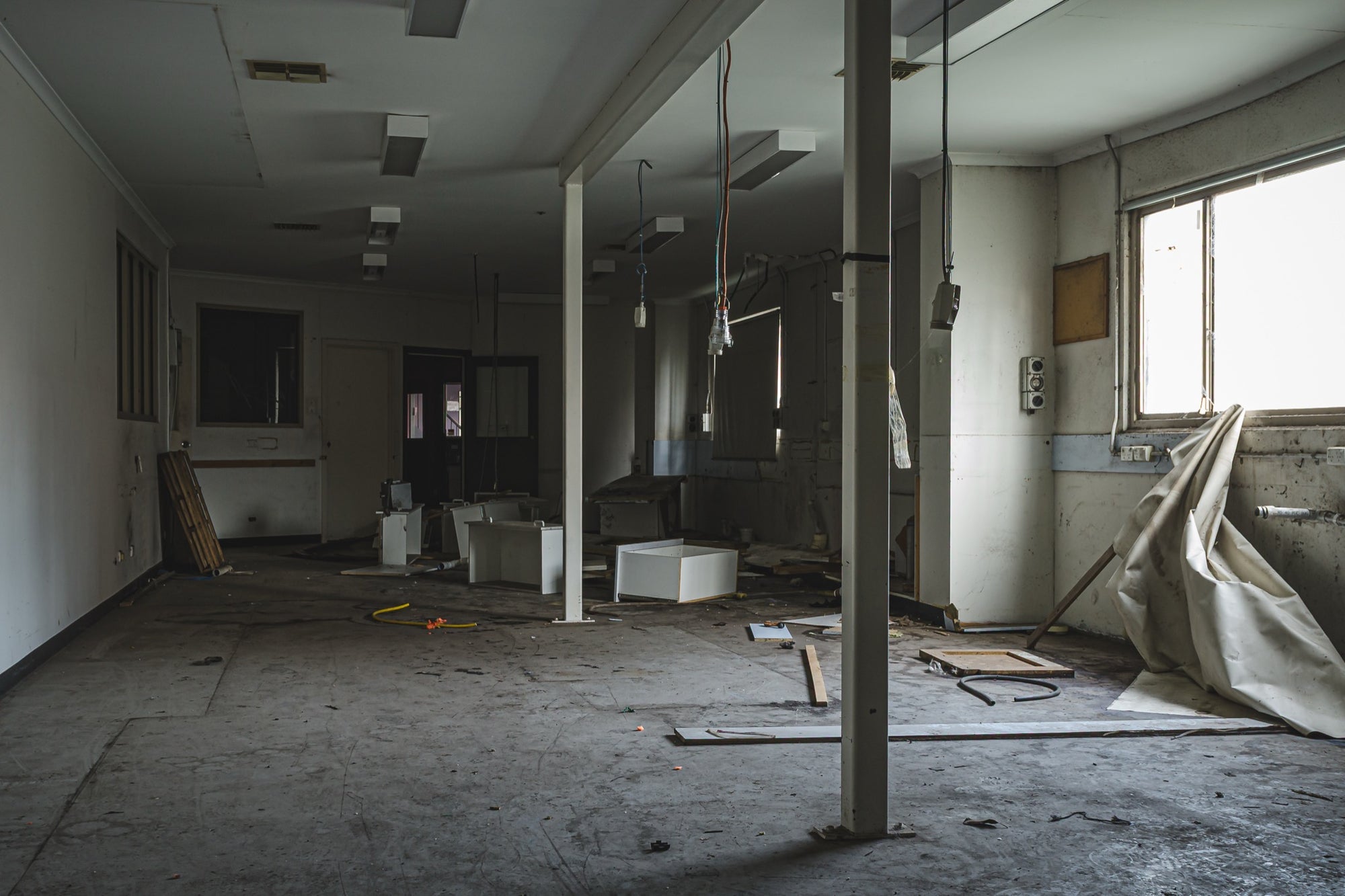 Stripped interior room at the former Bradmill factory in Yarraville, with a white support column, scattered cabinetry panels and cables on a bare concrete floor, and daylight entering from a window on the right.