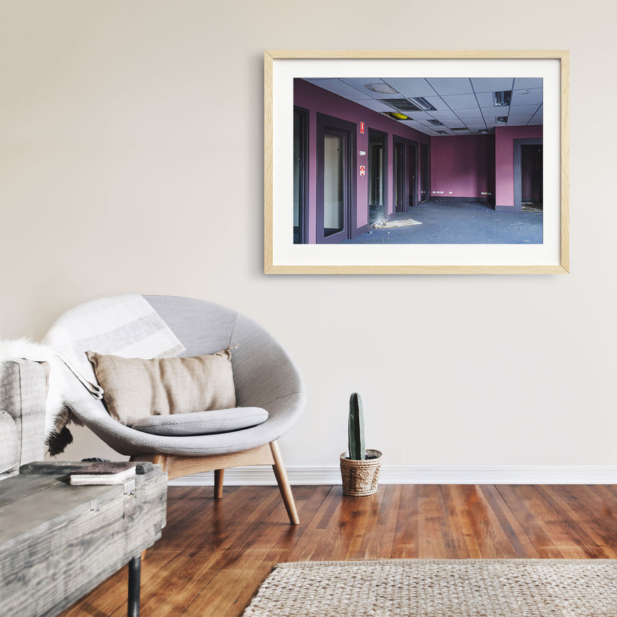 Empty office corridor at the former Bradmill factory in Yarraville, with mauve walls, grey carpet, glazed timber-framed doorways, a damaged suspended ceiling, and a red fire safety sign on the wall.