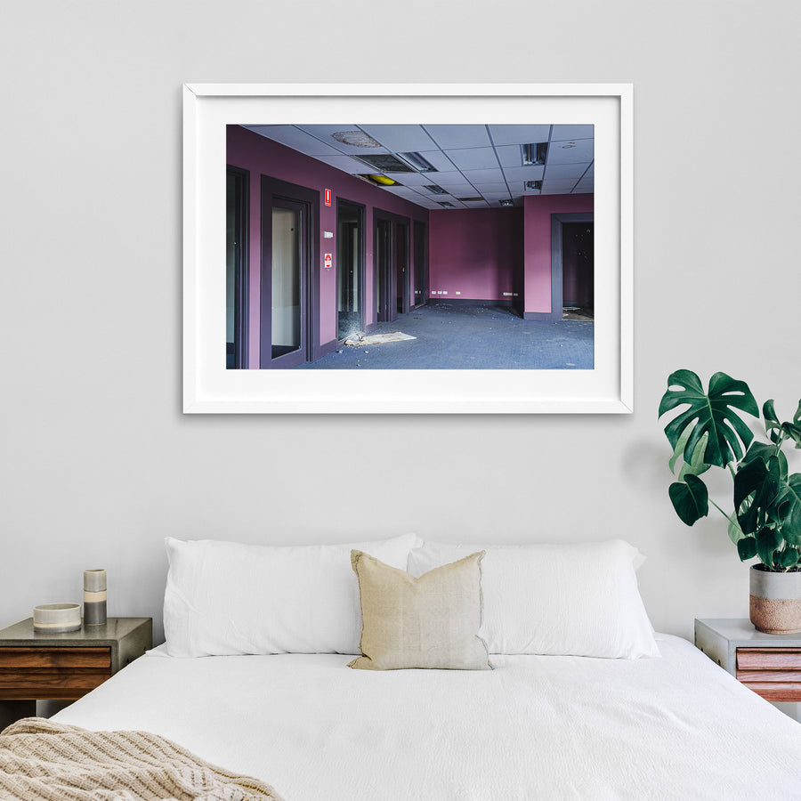 Empty office corridor at the former Bradmill factory in Yarraville, with mauve walls, grey carpet, glazed timber-framed doorways, a damaged suspended ceiling, and a red fire safety sign on the wall.