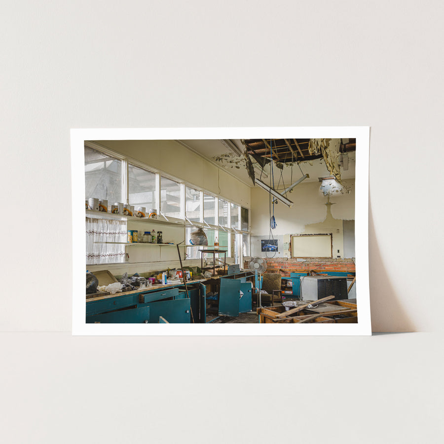 Derelict staff room interior at the former Bradmill factory in Yarraville, with rows of blue cabinets, scattered debris on benchtops, a collapsed ceiling exposing timber battens, and tins on a high shelf above tall windows.