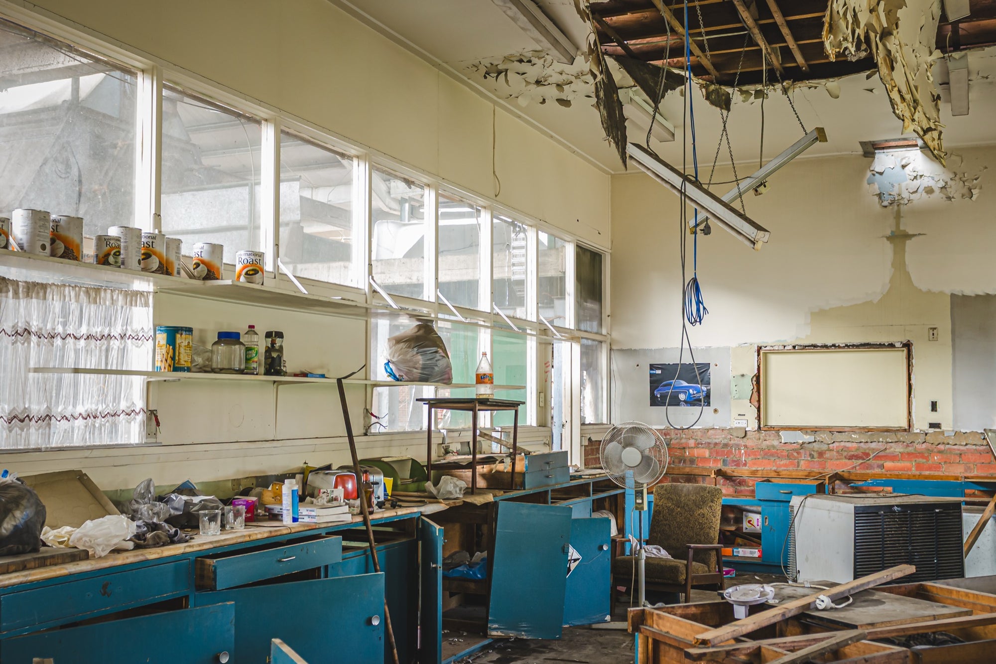 Derelict staff room interior at the former Bradmill factory in Yarraville, with rows of blue cabinets, scattered debris on benchtops, a collapsed ceiling exposing timber battens, and tins on a high shelf above tall windows.