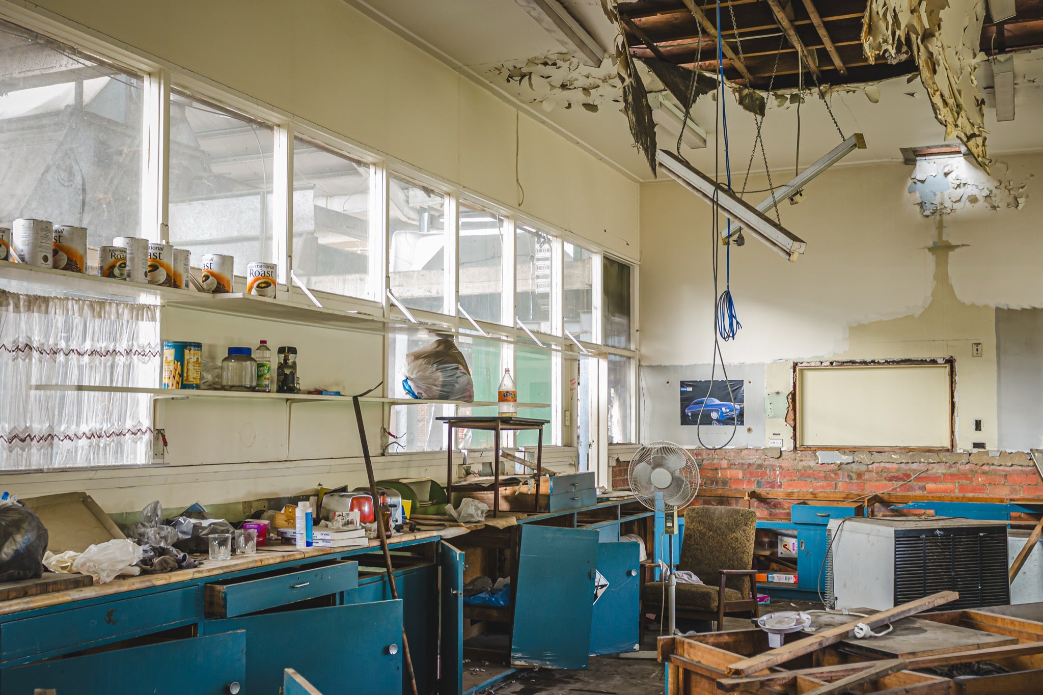Derelict staff room interior at the former Bradmill factory in Yarraville, with rows of blue cabinets, scattered debris on benchtops, a collapsed ceiling exposing timber battens, and tins on a high shelf above tall windows.