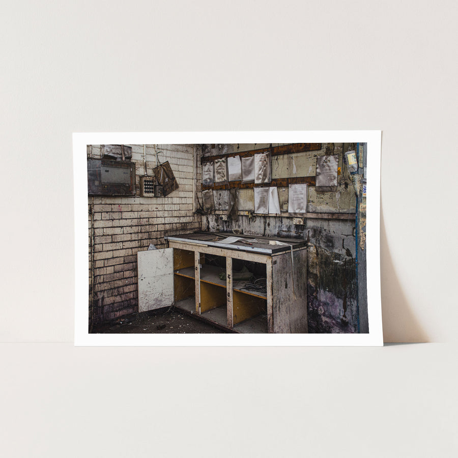 A worn workbench with a laminate top, open lower cabinet and detached door, small tools on the surface and faded documents pinned to the brick wall behind, photographed at the former Davies Coop / Bradmill factory in Yarraville.