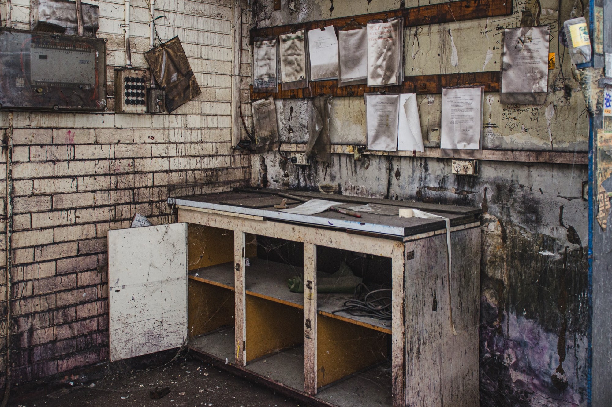 A worn workbench with a laminate top, open lower cabinet and detached door, small tools on the surface and faded documents pinned to the brick wall behind, photographed at the former Davies Coop / Bradmill factory in Yarraville.
