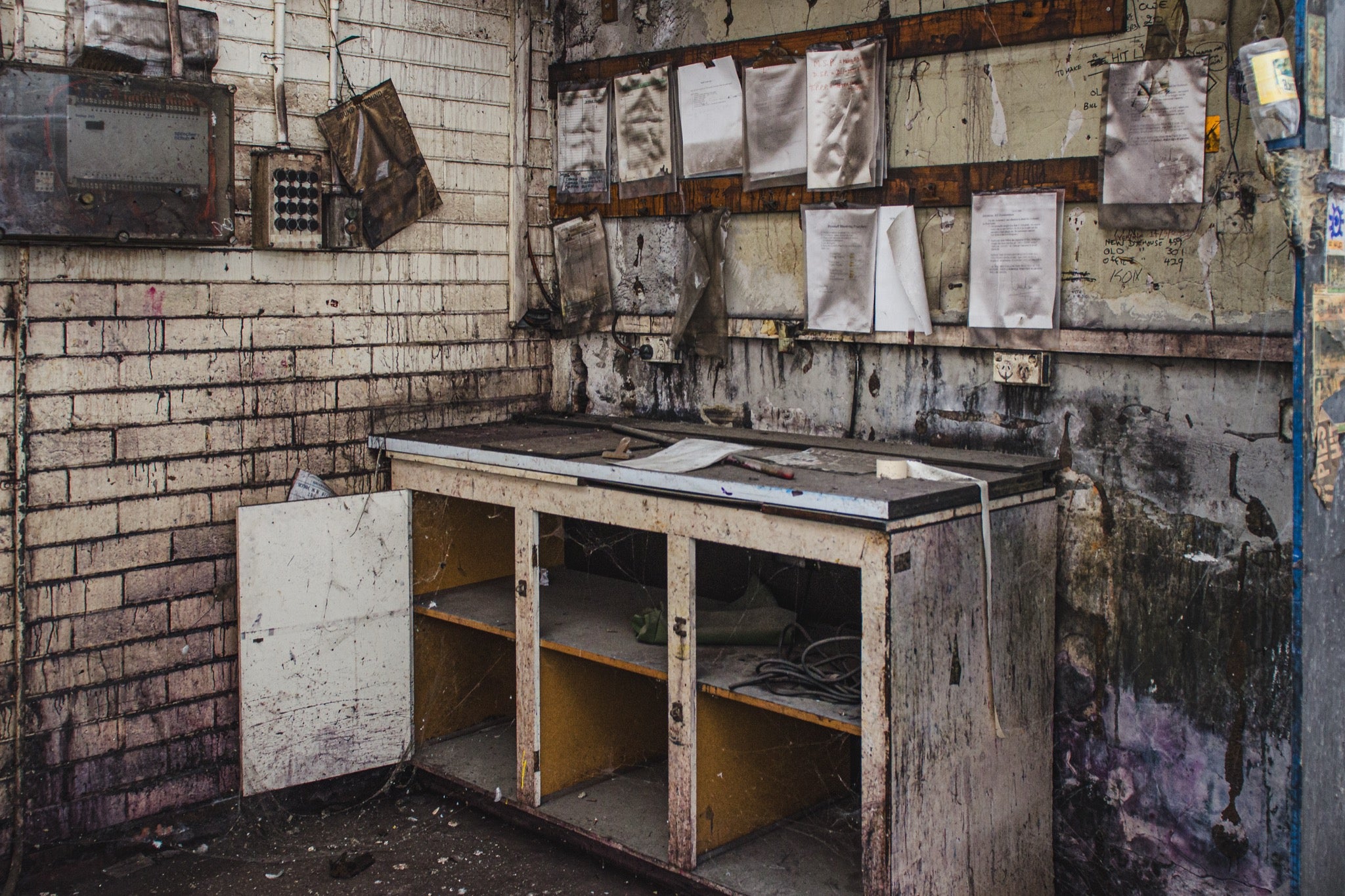A worn workbench with a laminate top, open lower cabinet and detached door, small tools on the surface and faded documents pinned to the brick wall behind, photographed at the former Davies Coop / Bradmill factory in Yarraville.