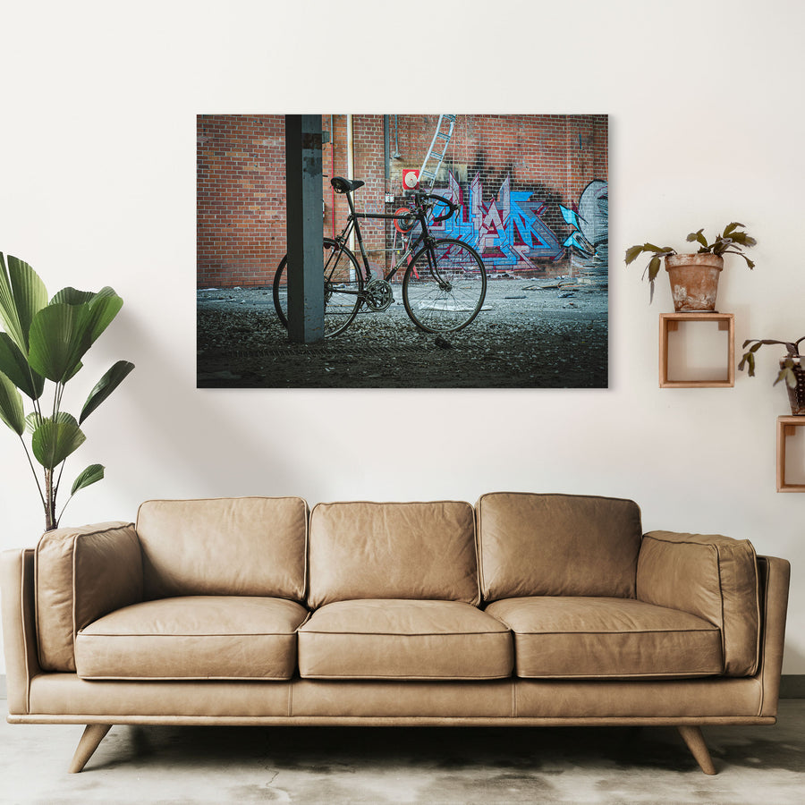 A worn black road bicycle leaning against a concrete pillar at the former Bradmill factory in Yarraville, with graffiti-covered brick wall, metal ladder, and red circular target visible behind it.