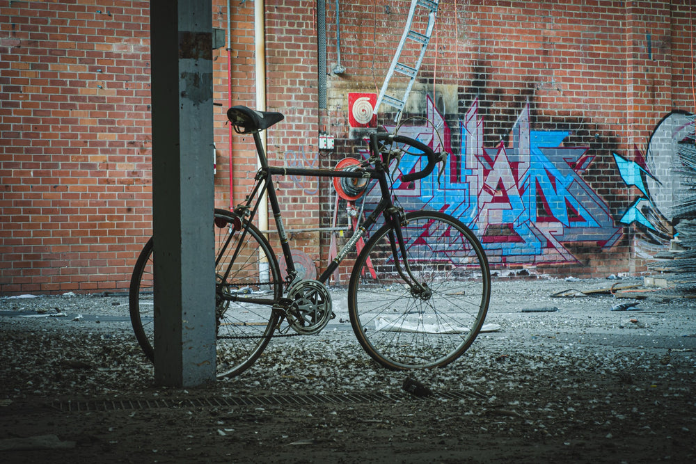 A worn black road bicycle leaning against a concrete pillar at the former Bradmill factory in Yarraville, with graffiti-covered brick wall, metal ladder, and red circular target visible behind it.
