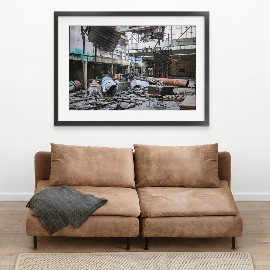 Fire-damaged manufacturing hall at the former Bradmill factory in Yarraville, with a collapsed roof, twisted metal sheeting, rusted cylindrical vessels, and a single chair on the debris-covered floor.