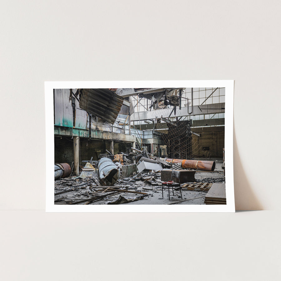 Fire-damaged manufacturing hall at the former Bradmill factory in Yarraville, with a collapsed roof, twisted metal sheeting, rusted cylindrical vessels, and a single chair on the debris-covered floor.