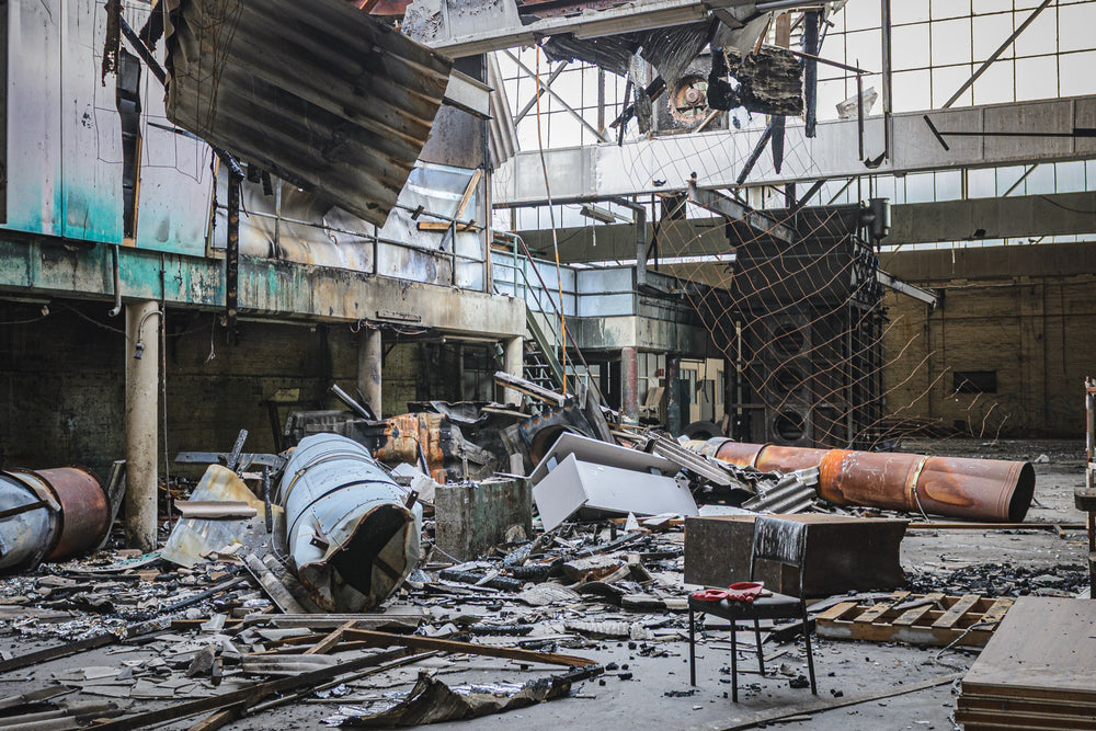 Fire-damaged manufacturing hall at the former Bradmill factory in Yarraville, with a collapsed roof, twisted metal sheeting, rusted cylindrical vessels, and a single chair on the debris-covered floor.