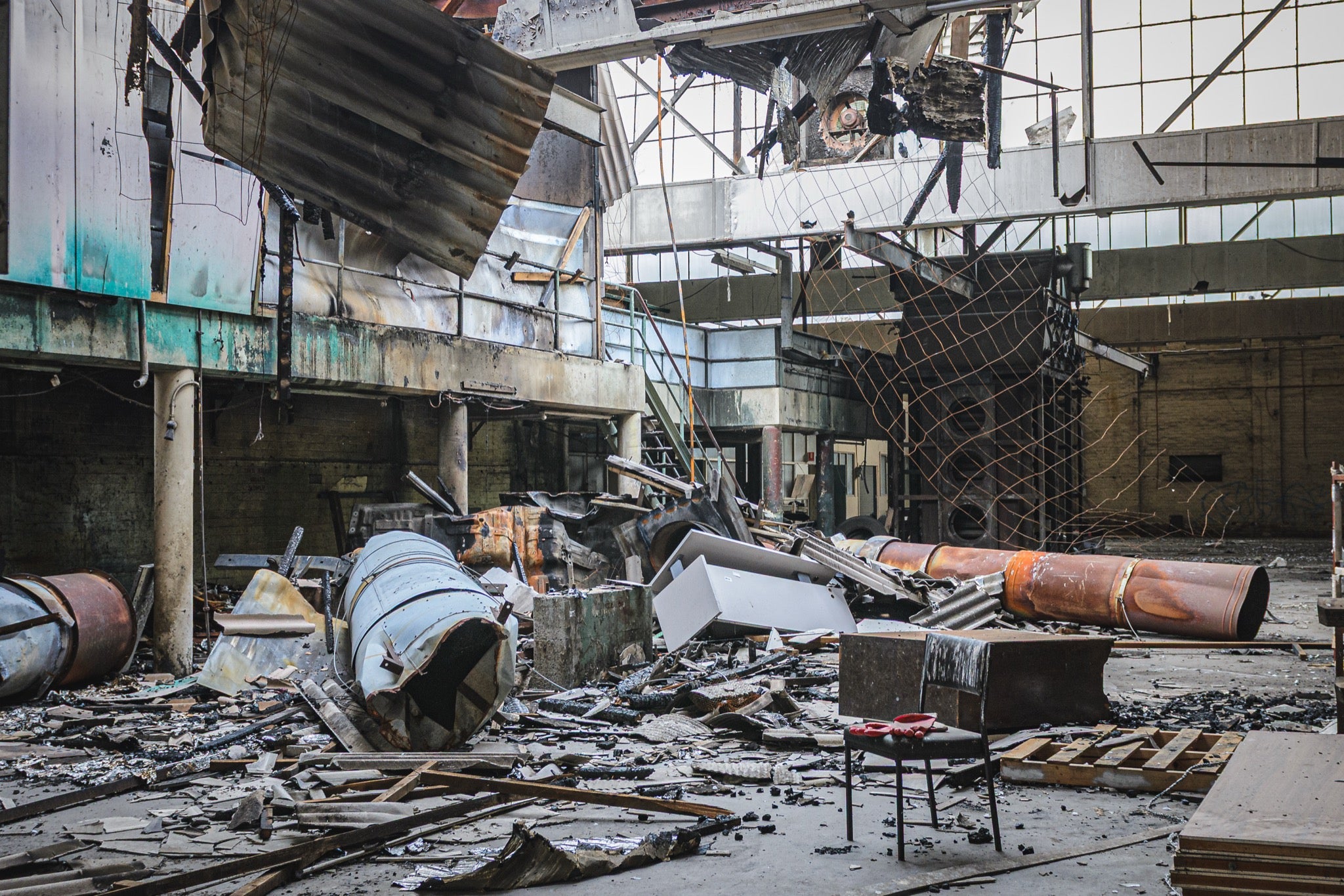Fire-damaged manufacturing hall at the former Bradmill factory in Yarraville, with a collapsed roof, twisted metal sheeting, rusted cylindrical vessels, and a single chair on the debris-covered floor.