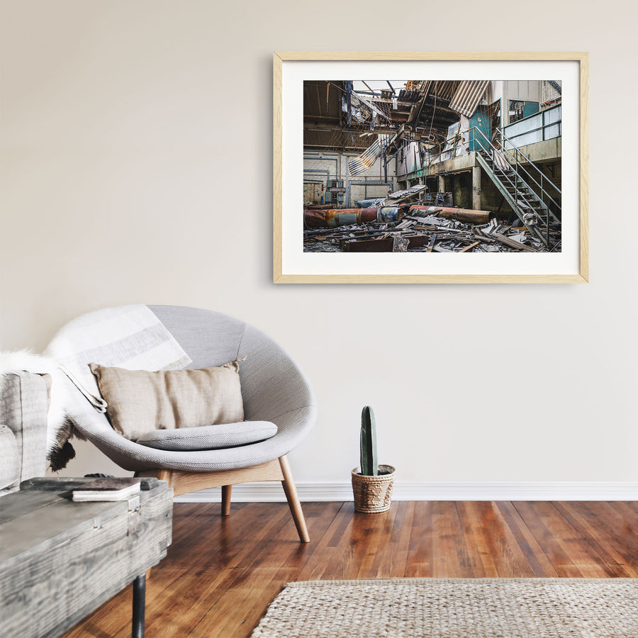 Collapsed corrugated roof sheeting, burnt timber and toppled cylindrical tanks covering the floor of a large industrial hall at the former Bradmill factory in Yarraville, with a steel staircase rising to a concrete mezzanine and old electrical boxes along a pale brick wall.