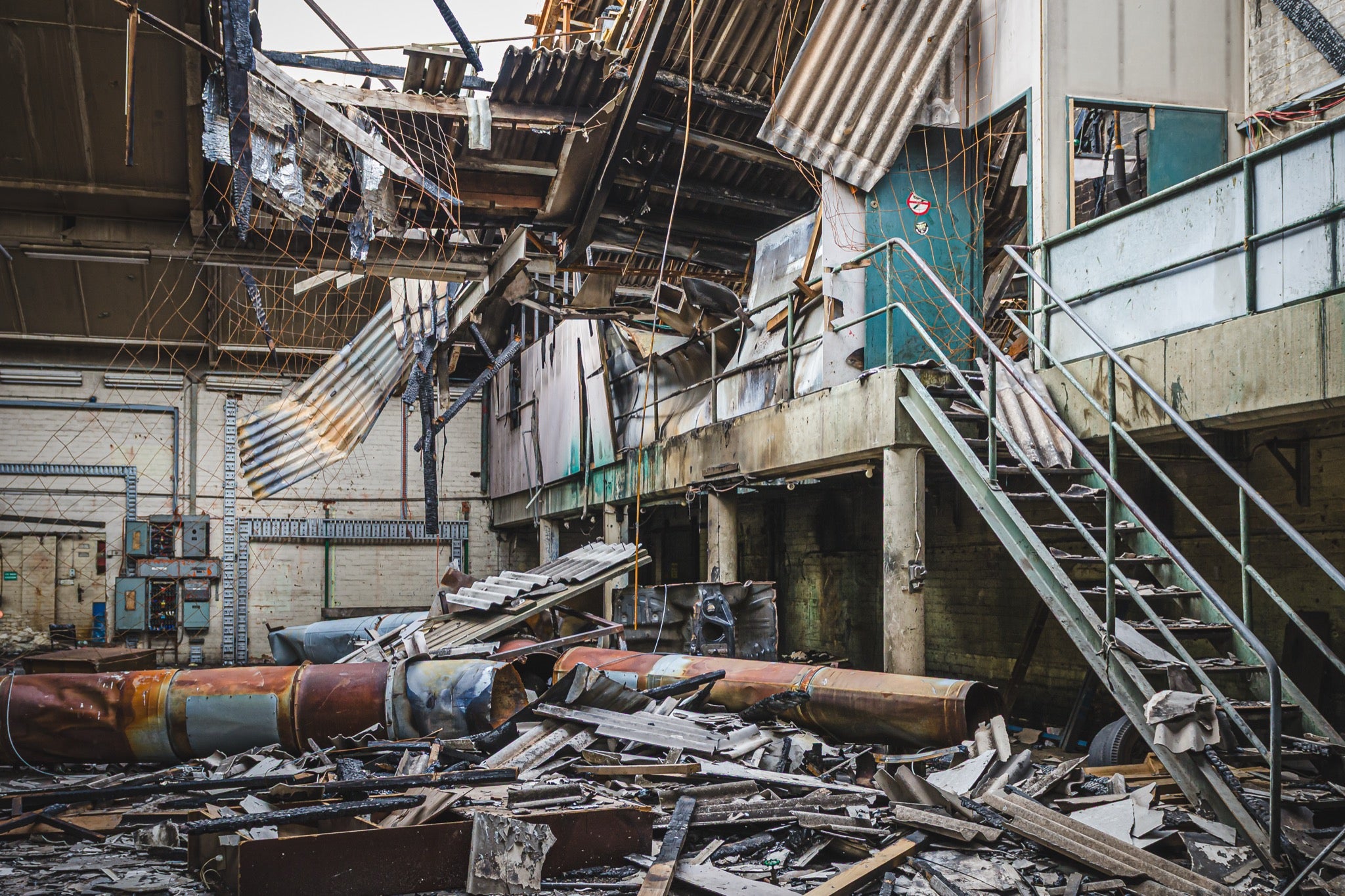 Collapsed corrugated roof sheeting, burnt timber and toppled cylindrical tanks covering the floor of a large industrial hall at the former Bradmill factory in Yarraville, with a steel staircase rising to a concrete mezzanine and old electrical boxes along a pale brick wall.