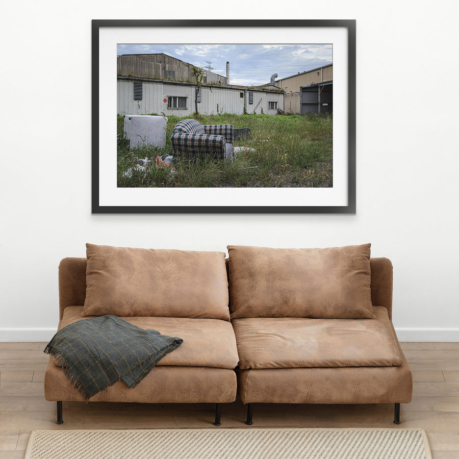 A patterned armchair sitting in long grass in front of metal-clad industrial sheds and a tall chimney at the former Davies Coop / Bradmill factory, Yarraville.