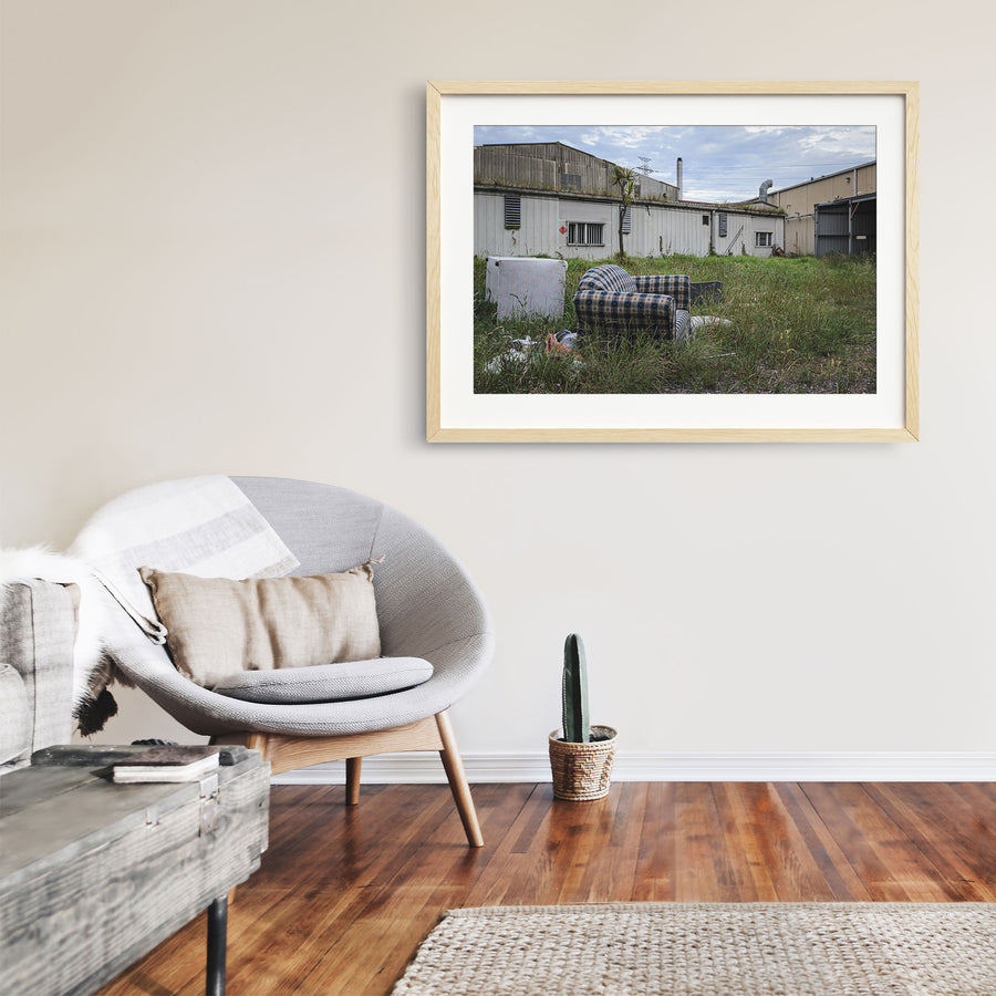 A patterned armchair sitting in long grass in front of metal-clad industrial sheds and a tall chimney at the former Davies Coop / Bradmill factory, Yarraville.
