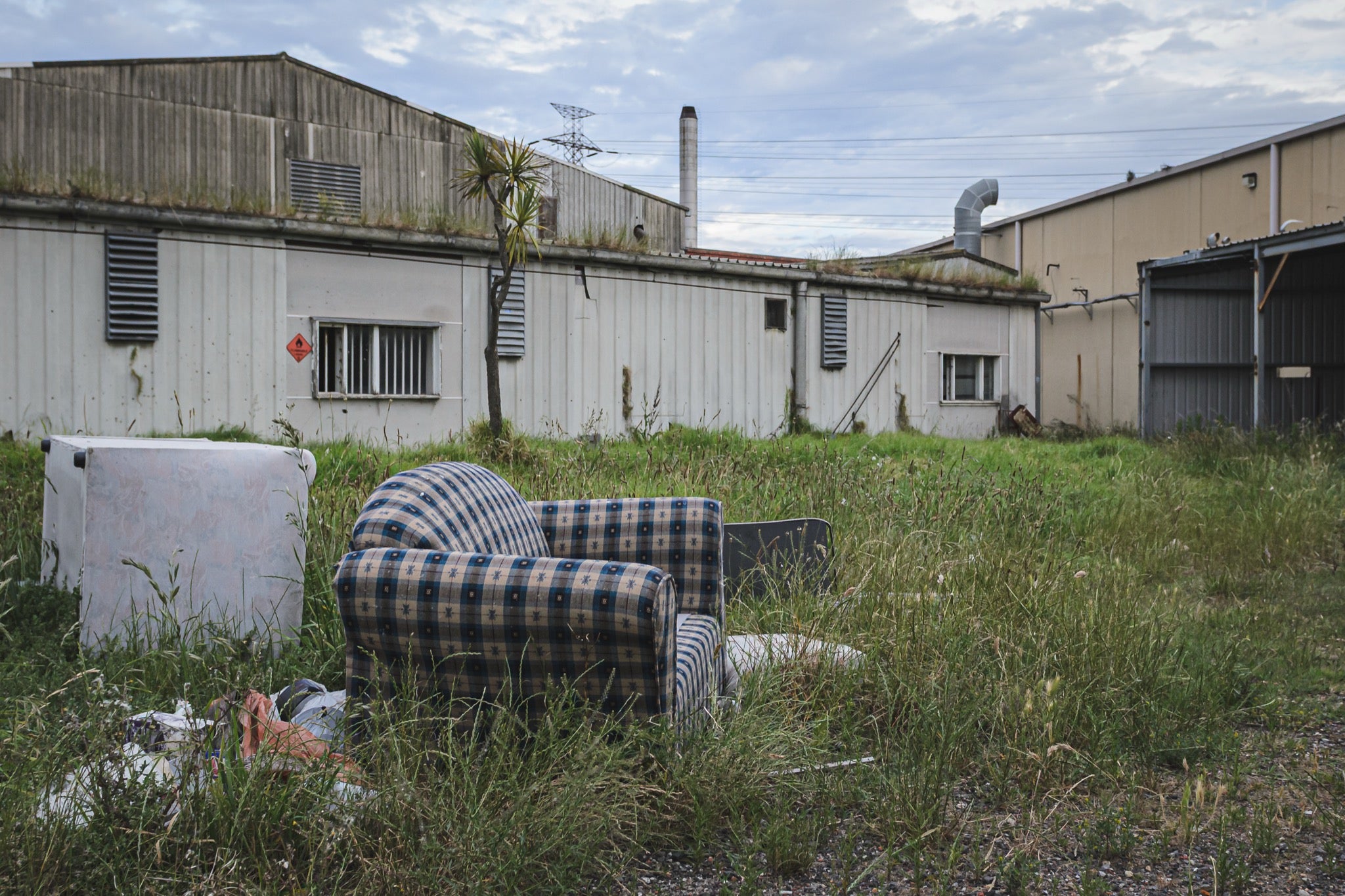 A patterned armchair sitting in long grass in front of metal-clad industrial sheds and a tall chimney at the former Davies Coop / Bradmill factory, Yarraville.