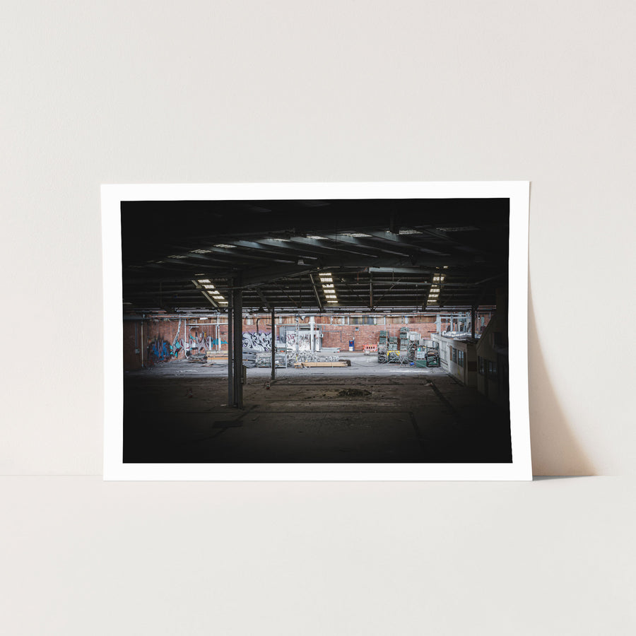 Empty warehouse interior at the former Bradmill factory in Yarraville, showing a steel-framed roof with skylights, graffiti-covered brick walls, and pallets and stacked timber across the concrete floor.