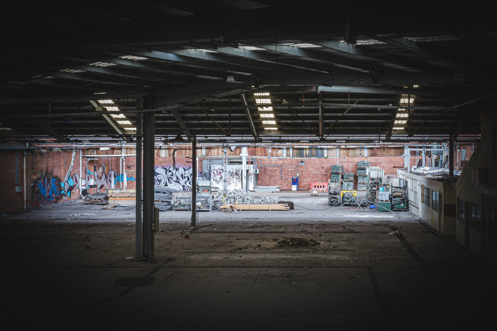 Empty warehouse interior at the former Bradmill factory in Yarraville, showing a steel-framed roof with skylights, graffiti-covered brick walls, and pallets and stacked timber across the concrete floor.