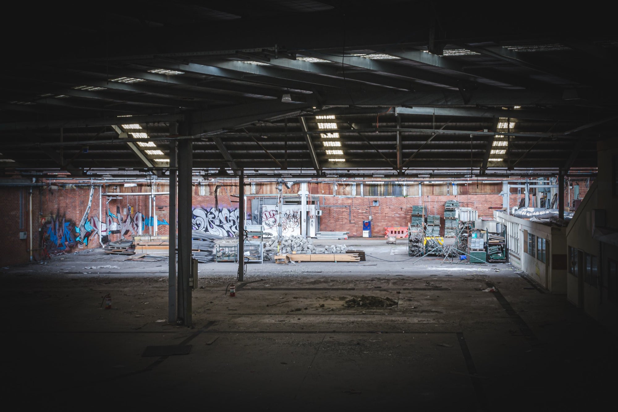 Empty warehouse interior at the former Bradmill factory in Yarraville, showing a steel-framed roof with skylights, graffiti-covered brick walls, and pallets and stacked timber across the concrete floor.
