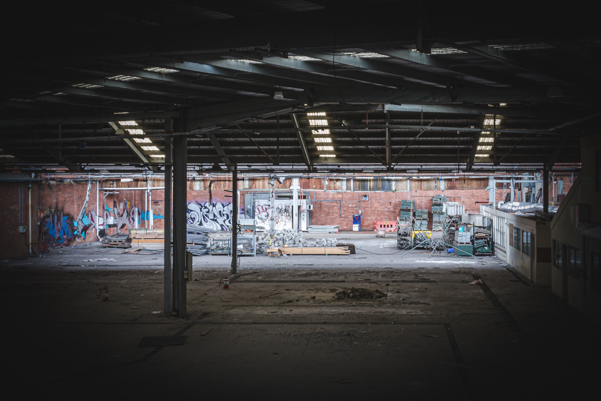 Empty warehouse interior at the former Bradmill factory in Yarraville, showing a steel-framed roof with skylights, graffiti-covered brick walls, and pallets and stacked timber across the concrete floor.