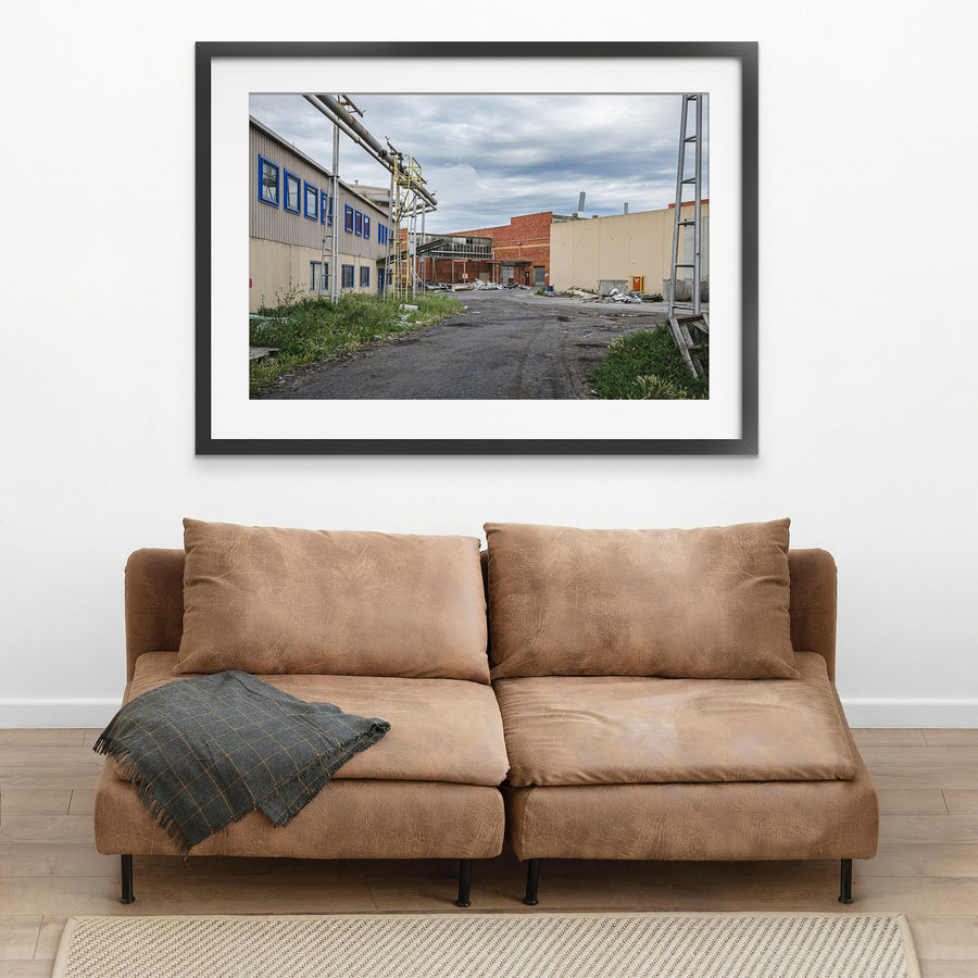 A gravel laneway between a clad industrial building with blue-framed windows and a brick warehouse with a chimney at the former Bradmill factory, Yarraville, with steel pipe gantries and a yellow caged ladder visible to one side.