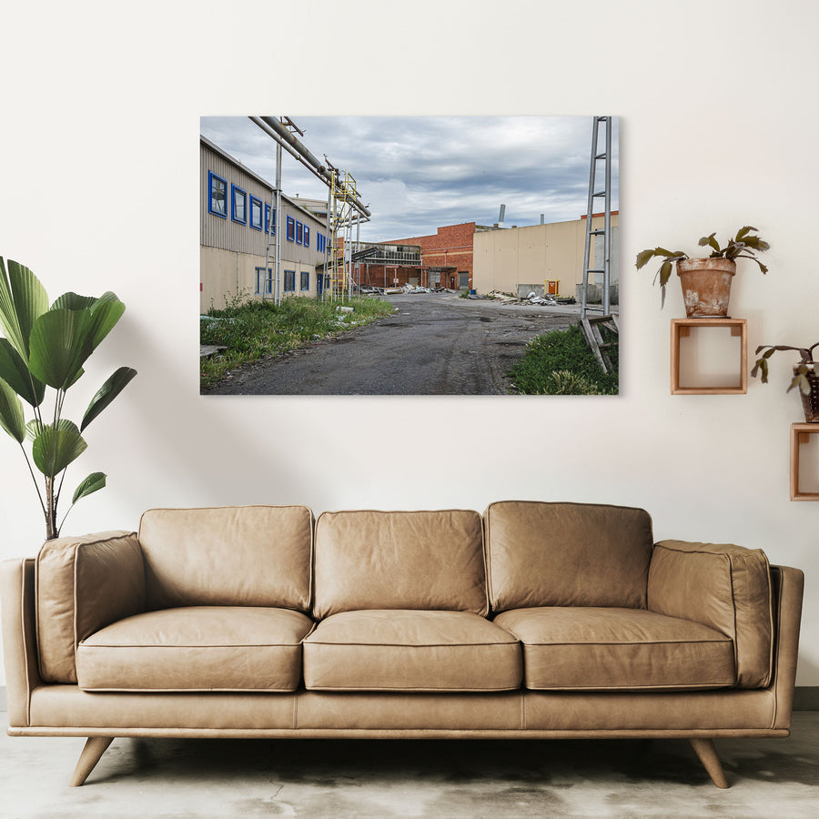 A gravel laneway between a clad industrial building with blue-framed windows and a brick warehouse with a chimney at the former Bradmill factory, Yarraville, with steel pipe gantries and a yellow caged ladder visible to one side.