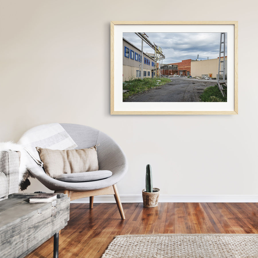 A gravel laneway between a clad industrial building with blue-framed windows and a brick warehouse with a chimney at the former Bradmill factory, Yarraville, with steel pipe gantries and a yellow caged ladder visible to one side.