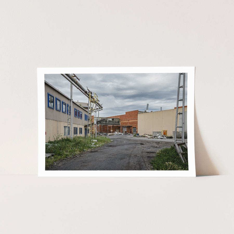 A gravel laneway between a clad industrial building with blue-framed windows and a brick warehouse with a chimney at the former Bradmill factory, Yarraville, with steel pipe gantries and a yellow caged ladder visible to one side.