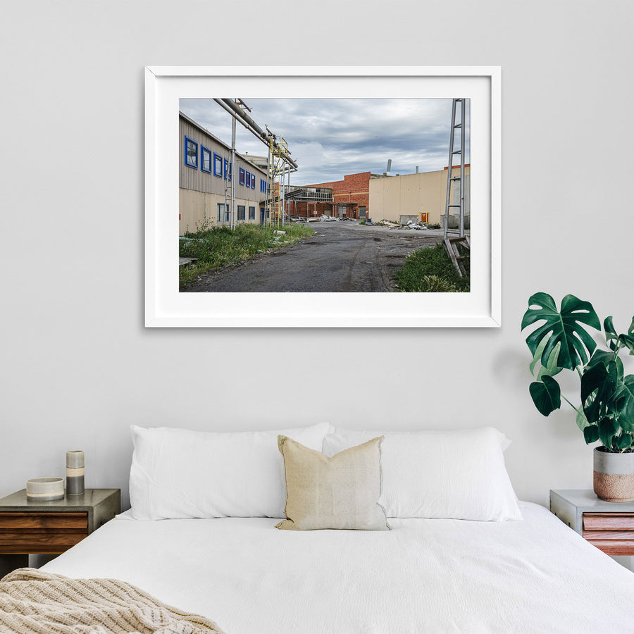 A gravel laneway between a clad industrial building with blue-framed windows and a brick warehouse with a chimney at the former Bradmill factory, Yarraville, with steel pipe gantries and a yellow caged ladder visible to one side.