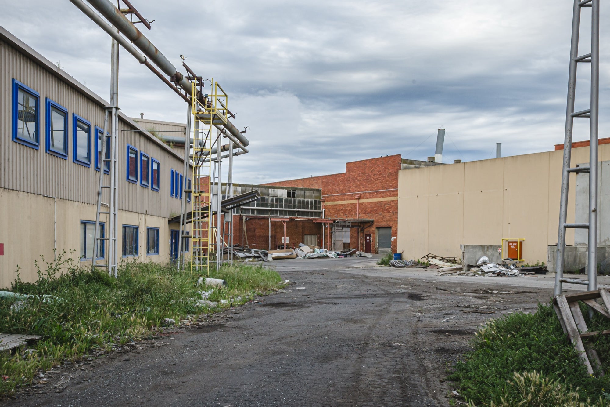 A gravel laneway between a clad industrial building with blue-framed windows and a brick warehouse with a chimney at the former Bradmill factory, Yarraville, with steel pipe gantries and a yellow caged ladder visible to one side.