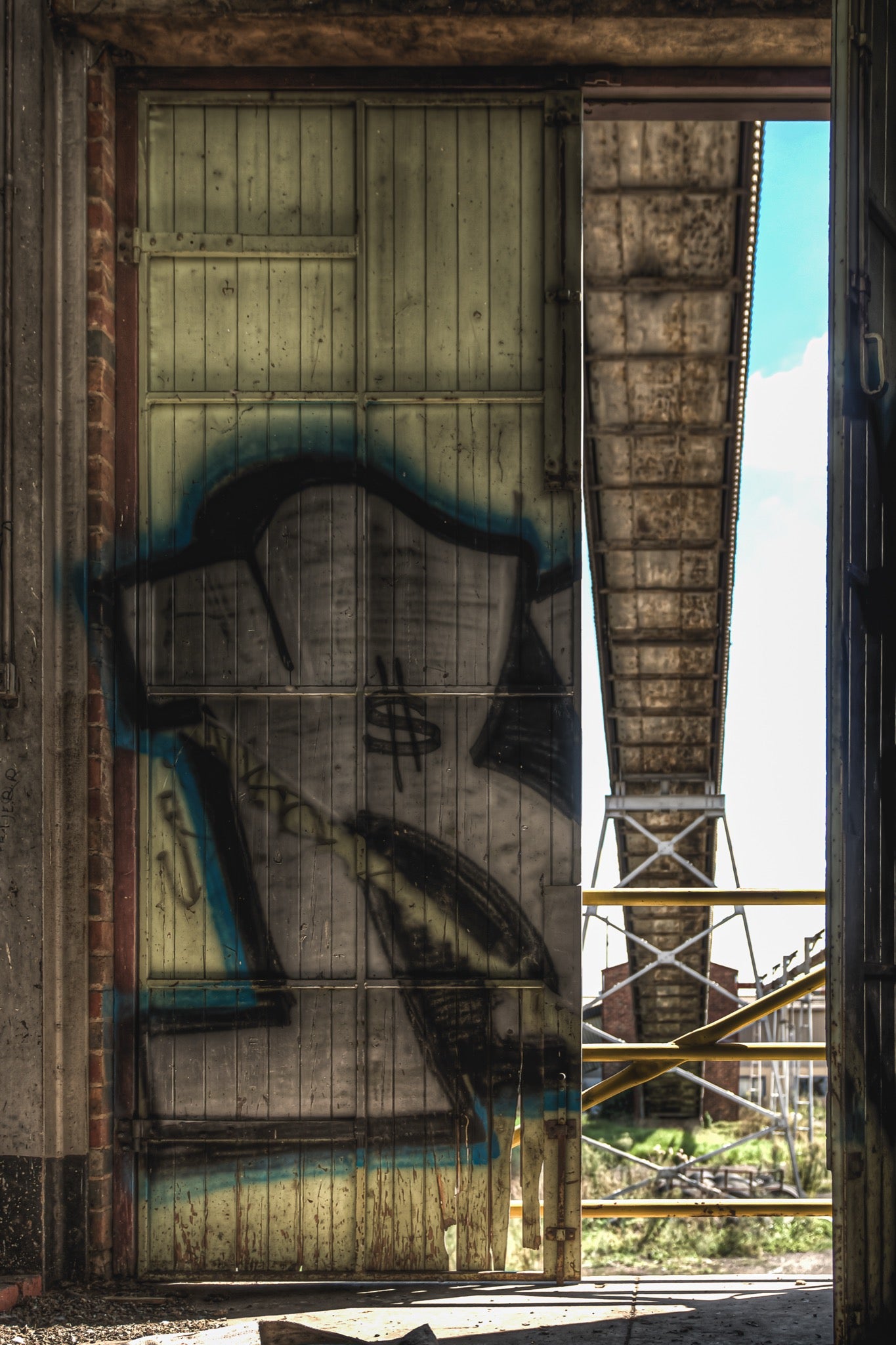 A rust-stained timber sliding door covered in spray-painted graffiti lettering at the former Bradmill factory in Yarraville, with a steel girder structure and yellow scaffolding visible through the gap beside it.