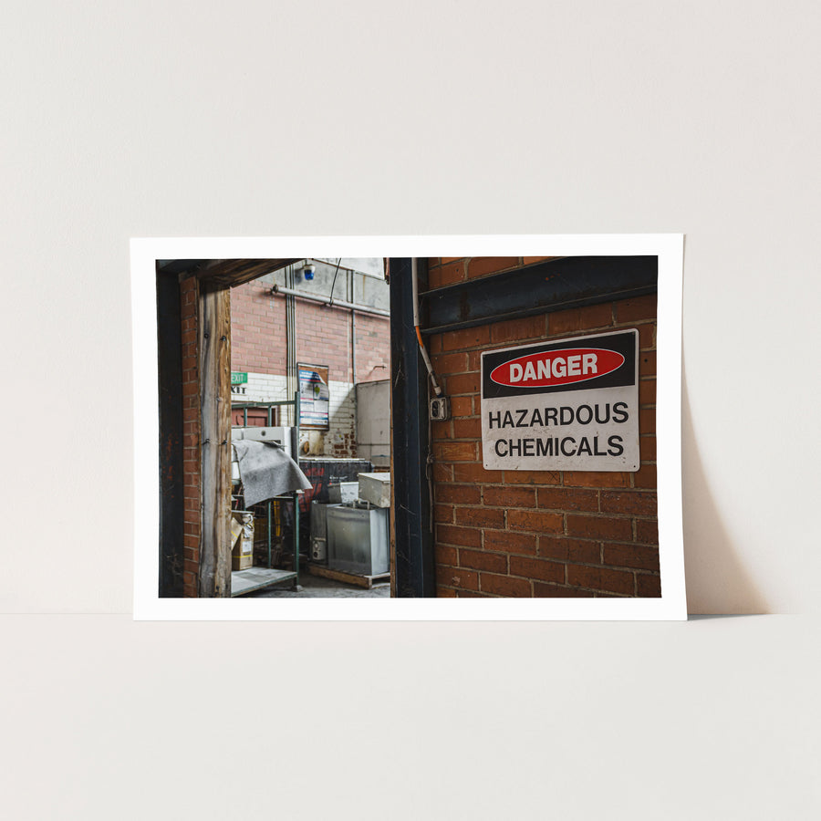 A weathered "Danger Hazardous Chemicals" sign on a brick wall beside an open timber doorway at the former Davies Coop / Bradmill factory in Yarraville, with metal cabinets, storage racks and an illuminated exit sign visible inside.