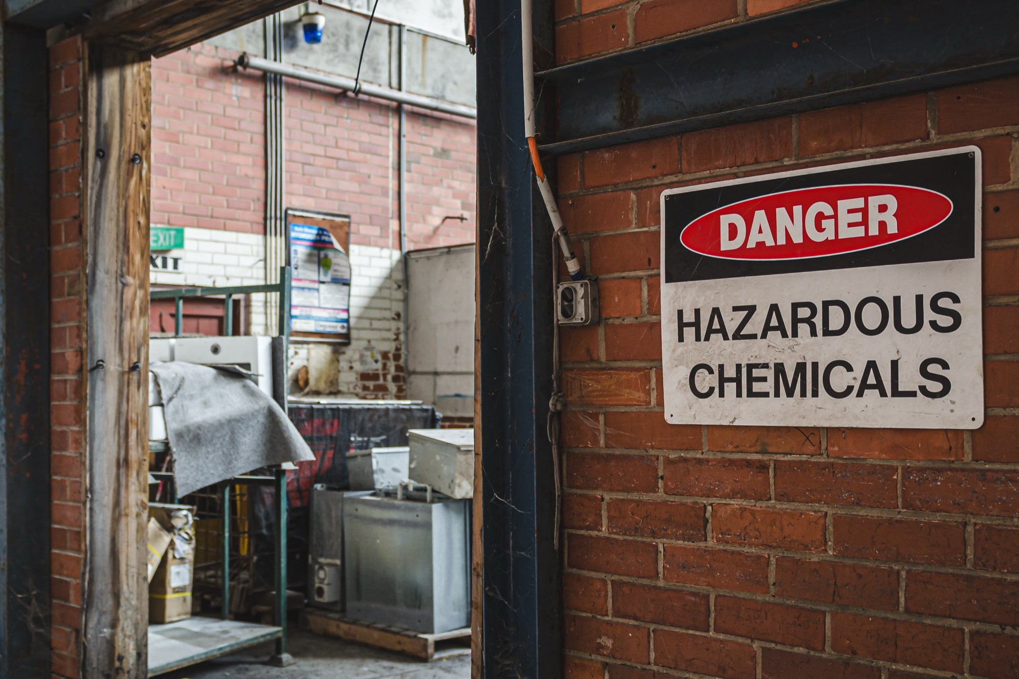 A weathered "Danger Hazardous Chemicals" sign on a brick wall beside an open timber doorway at the former Davies Coop / Bradmill factory in Yarraville, with metal cabinets, storage racks and an illuminated exit sign visible inside.