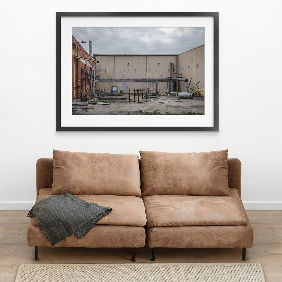 A concrete yard at the former Bradmill factory in Yarraville, enclosed by rendered walls and a brick building, with disused ducting, metal drums, a galvanised tub and severed pipework left across the slab under an overcast sky.