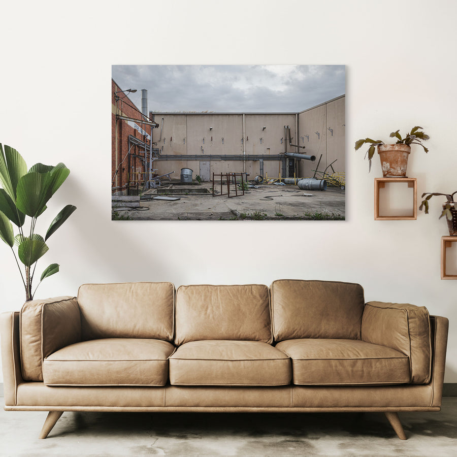 A concrete yard at the former Bradmill factory in Yarraville, enclosed by rendered walls and a brick building, with disused ducting, metal drums, a galvanised tub and severed pipework left across the slab under an overcast sky.