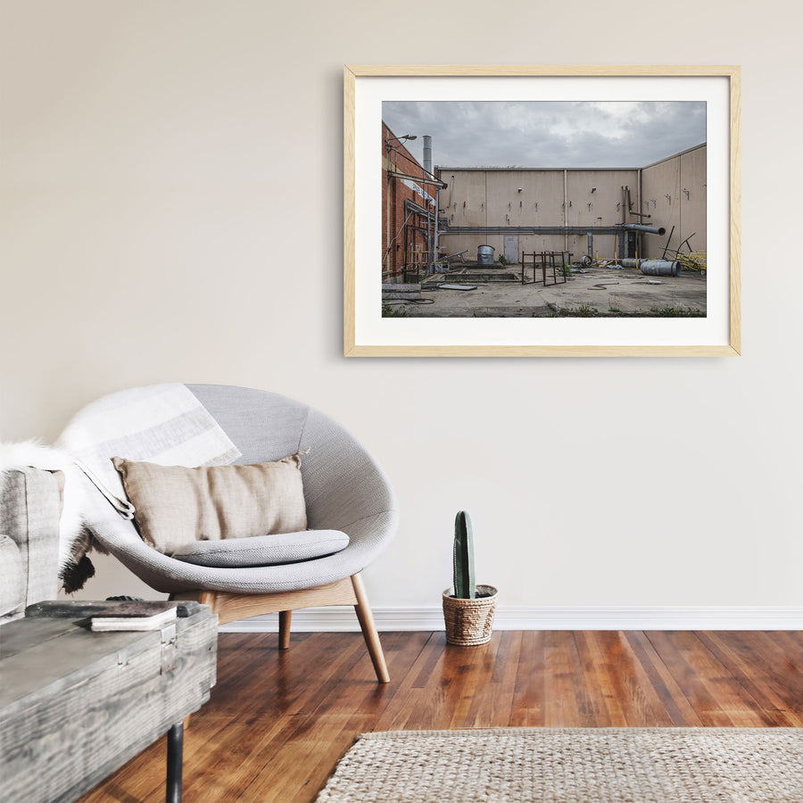 A concrete yard at the former Bradmill factory in Yarraville, enclosed by rendered walls and a brick building, with disused ducting, metal drums, a galvanised tub and severed pipework left across the slab under an overcast sky.