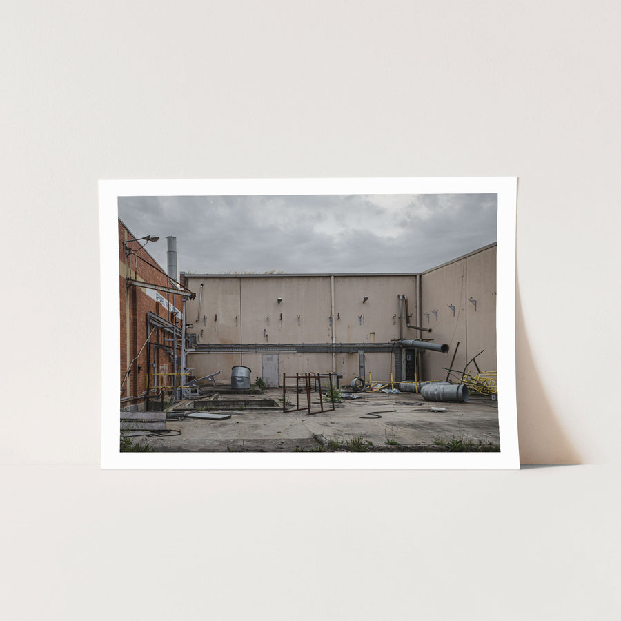 A concrete yard at the former Bradmill factory in Yarraville, enclosed by rendered walls and a brick building, with disused ducting, metal drums, a galvanised tub and severed pipework left across the slab under an overcast sky.