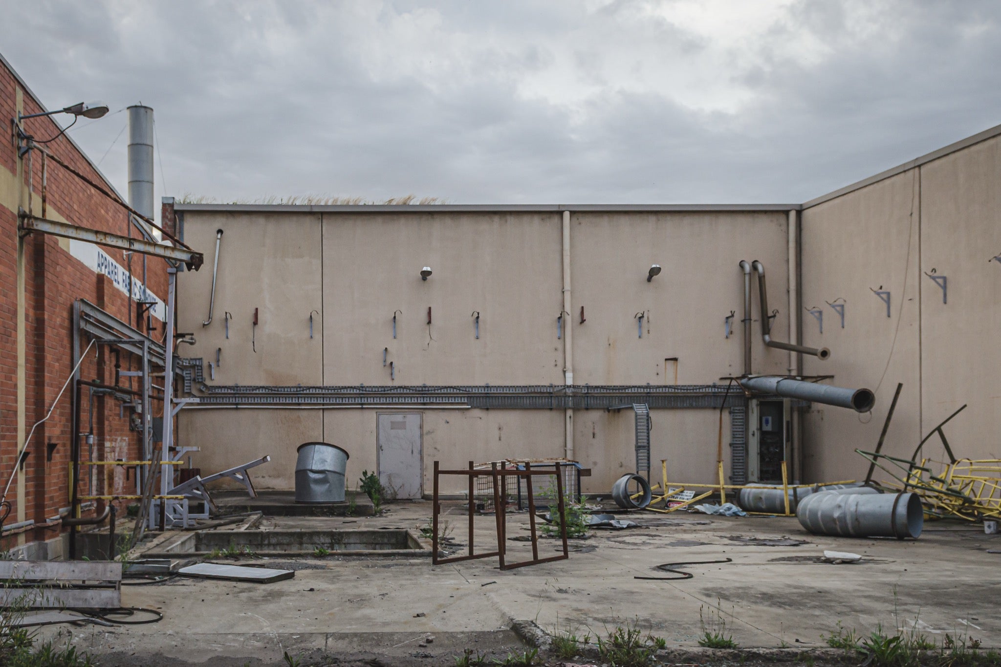 A concrete yard at the former Bradmill factory in Yarraville, enclosed by rendered walls and a brick building, with disused ducting, metal drums, a galvanised tub and severed pipework left across the slab under an overcast sky.