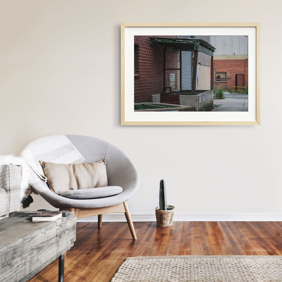 A green steel canopy over a concrete loading dock at the former Bradmill factory in Yarraville, with a closed corrugated roller door and a rusted metal bin below, a cracked yard and brick warehouse wall with a broken window beyond.