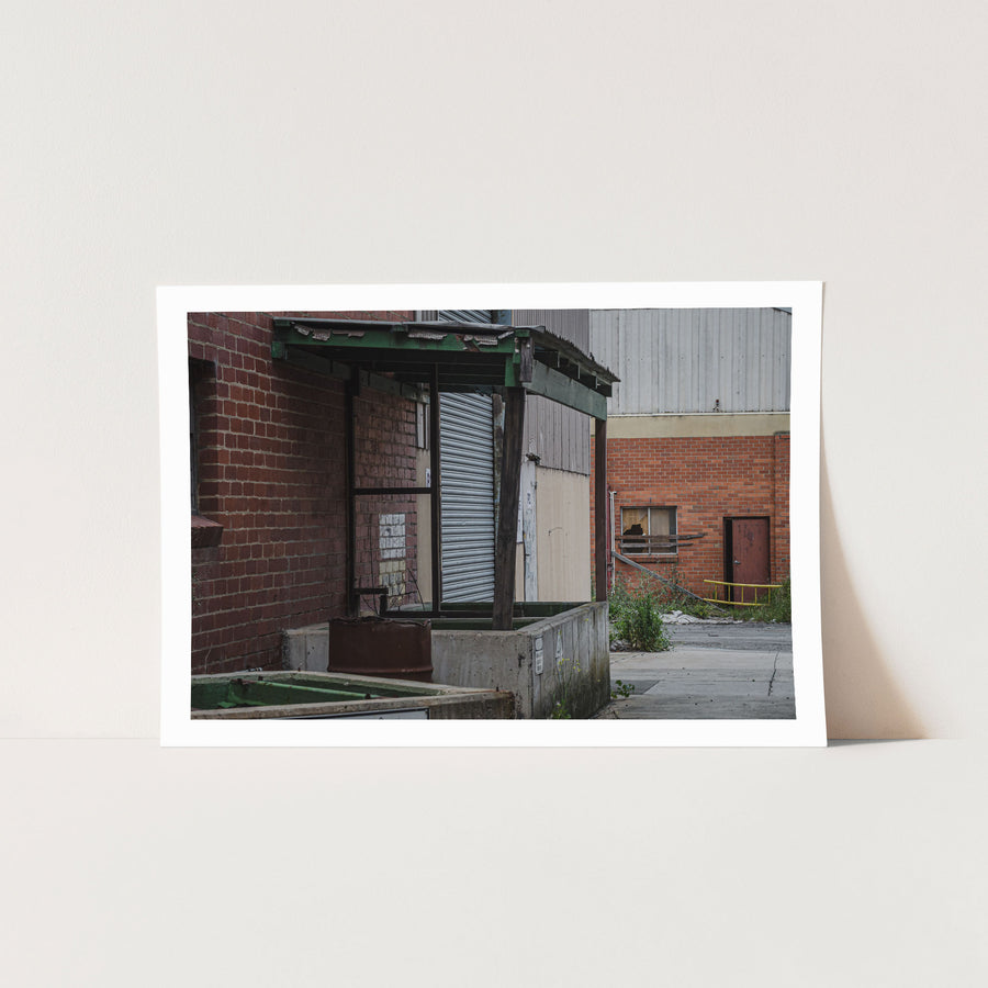 A green steel canopy over a concrete loading dock at the former Bradmill factory in Yarraville, with a closed corrugated roller door and a rusted metal bin below, a cracked yard and brick warehouse wall with a broken window beyond.