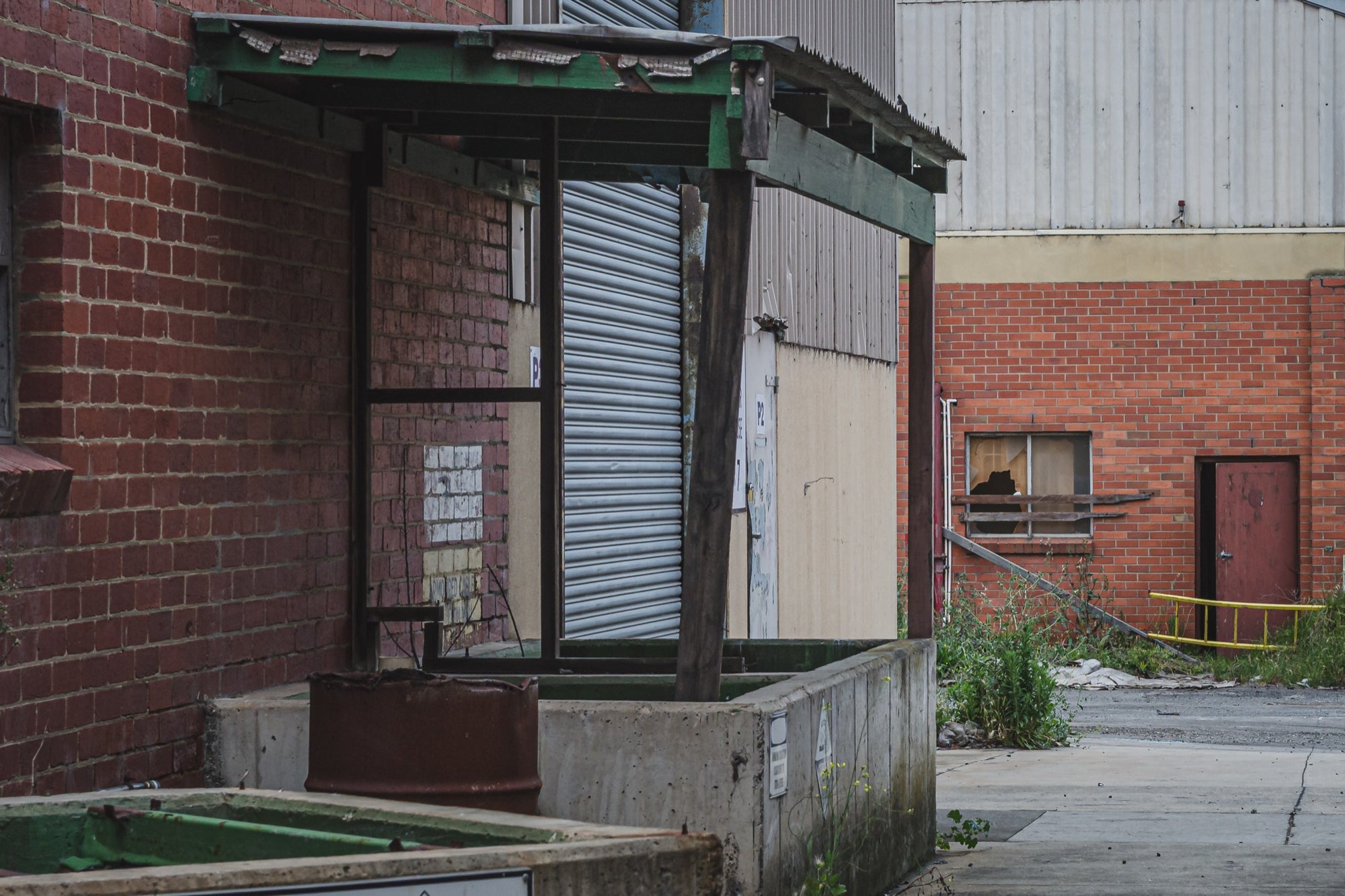 A green steel canopy over a concrete loading dock at the former Bradmill factory in Yarraville, with a closed corrugated roller door and a rusted metal bin below, a cracked yard and brick warehouse wall with a broken window beyond.