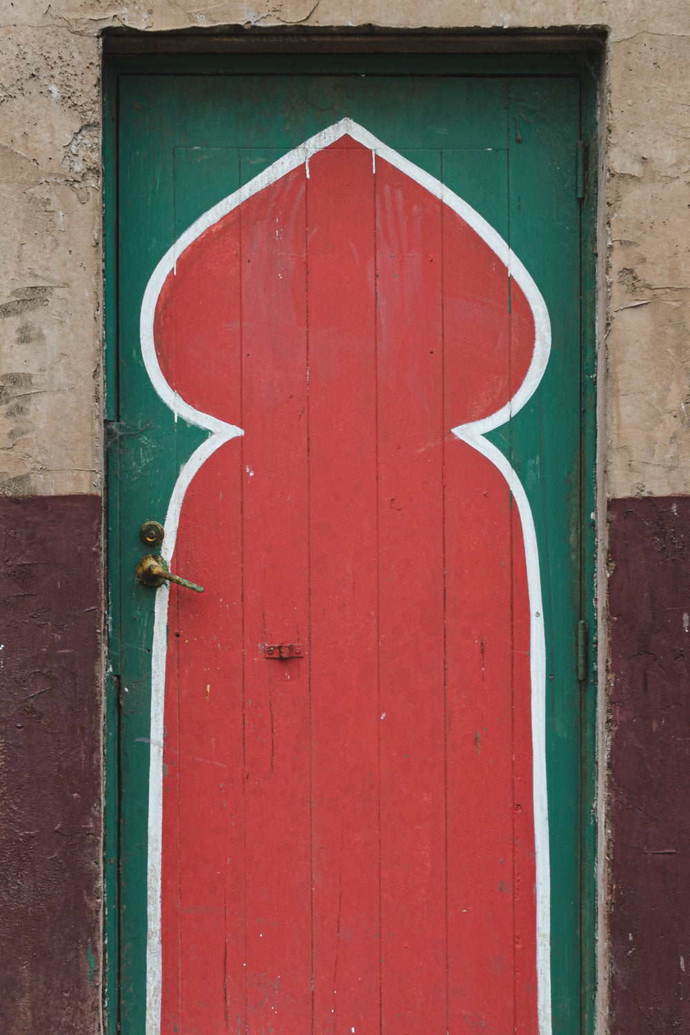 A painted timber door at the former Bradmill factory in Yarraville, with a tall red keyhole arch outlined in white on a green background, a worn brass lever handle on the left, and cracked render surrounding the frame.