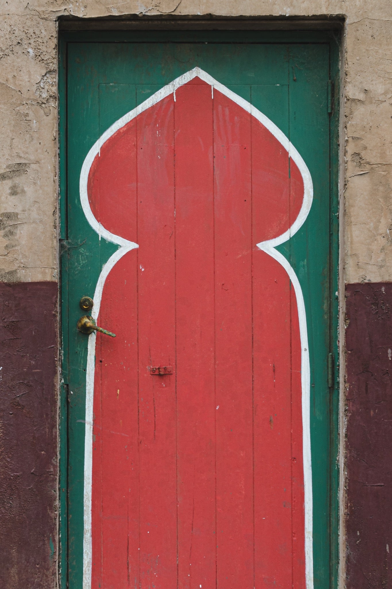 A painted timber door at the former Bradmill factory in Yarraville, with a tall red keyhole arch outlined in white on a green background, a worn brass lever handle on the left, and cracked render surrounding the frame.