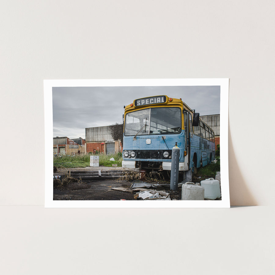 A weathered blue and yellow bus with a "Special" destination sign parked on a derelict overgrown lot at the former Bradmill factory in Yarraville, with corrugated iron and brick industrial buildings behind.