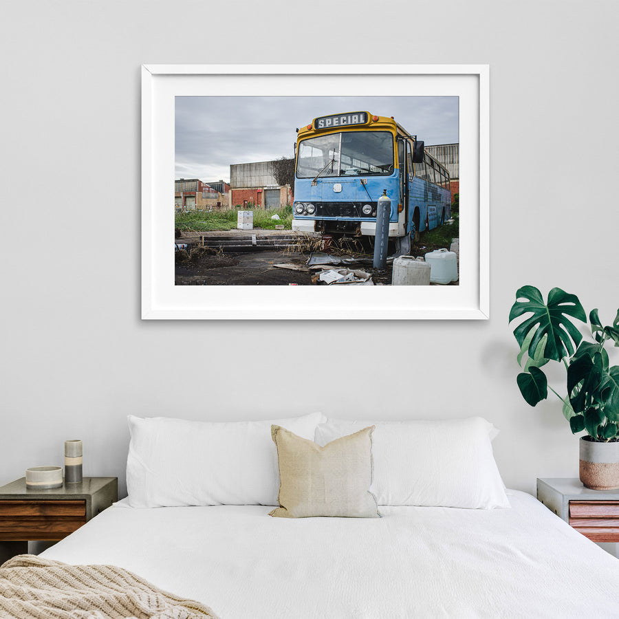 A weathered blue and yellow bus with a "Special" destination sign parked on a derelict overgrown lot at the former Bradmill factory in Yarraville, with corrugated iron and brick industrial buildings behind.