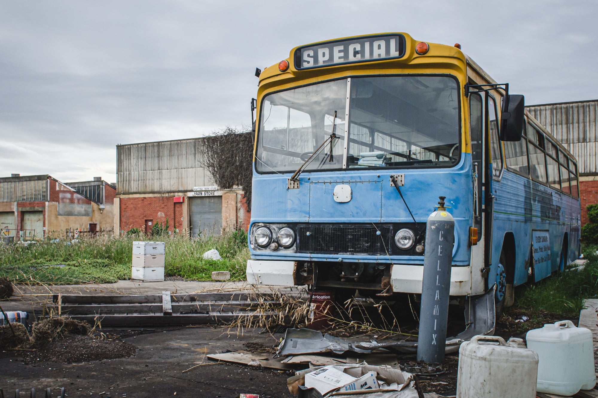 A weathered blue and yellow bus with a "Special" destination sign parked on a derelict overgrown lot at the former Bradmill factory in Yarraville, with corrugated iron and brick industrial buildings behind.