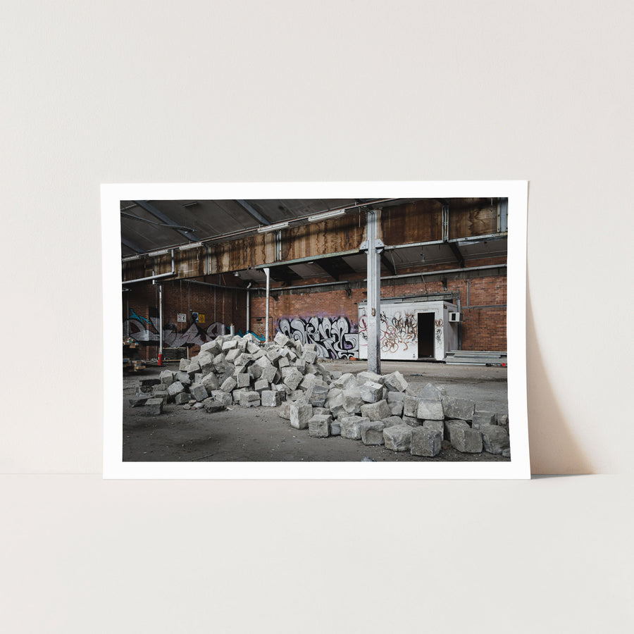 A pile of grey stone blocks on the dusty floor of an abandoned brick manufacturing building at the former Bradmill factory, Yarraville, with graffiti-covered walls and a rusting roof beam above.