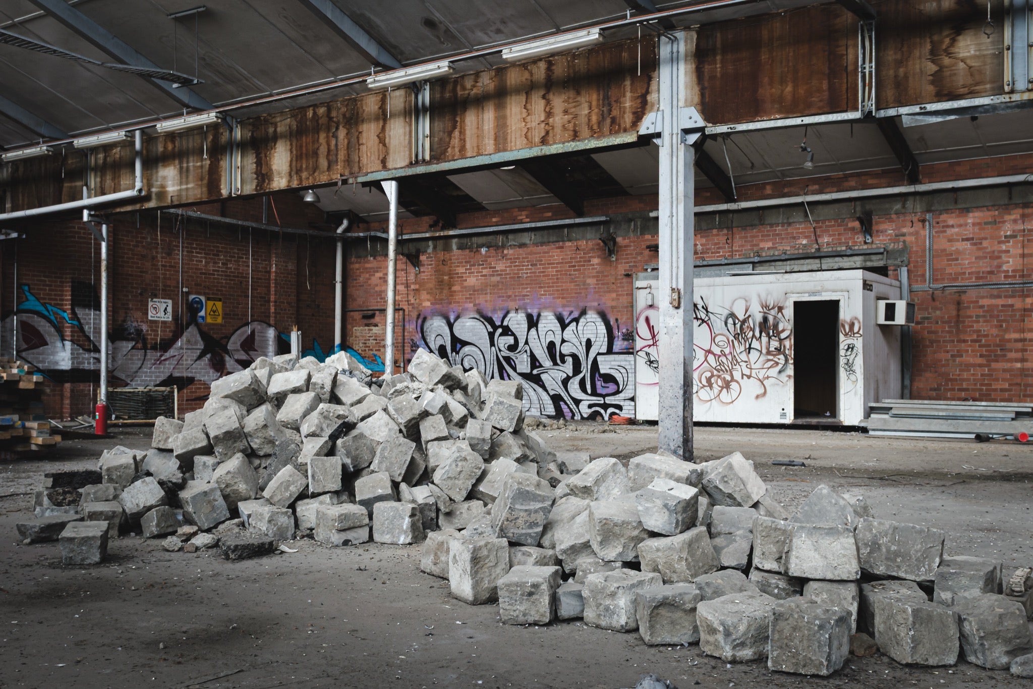 A pile of grey stone blocks on the dusty floor of an abandoned brick manufacturing building at the former Bradmill factory, Yarraville, with graffiti-covered walls and a rusting roof beam above.