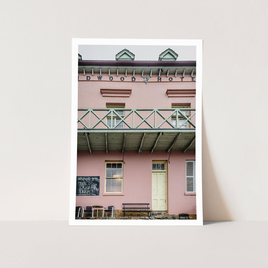 The pink rendered two-storey facade of Braidwood Hotel, with a timber balcony above a verandah, a chalkboard sign, and outdoor seating at street level.